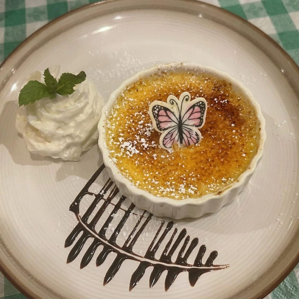 Crème brûlée with a butterfly decoration, whipped cream, and chocolate drizzle on a white plate.