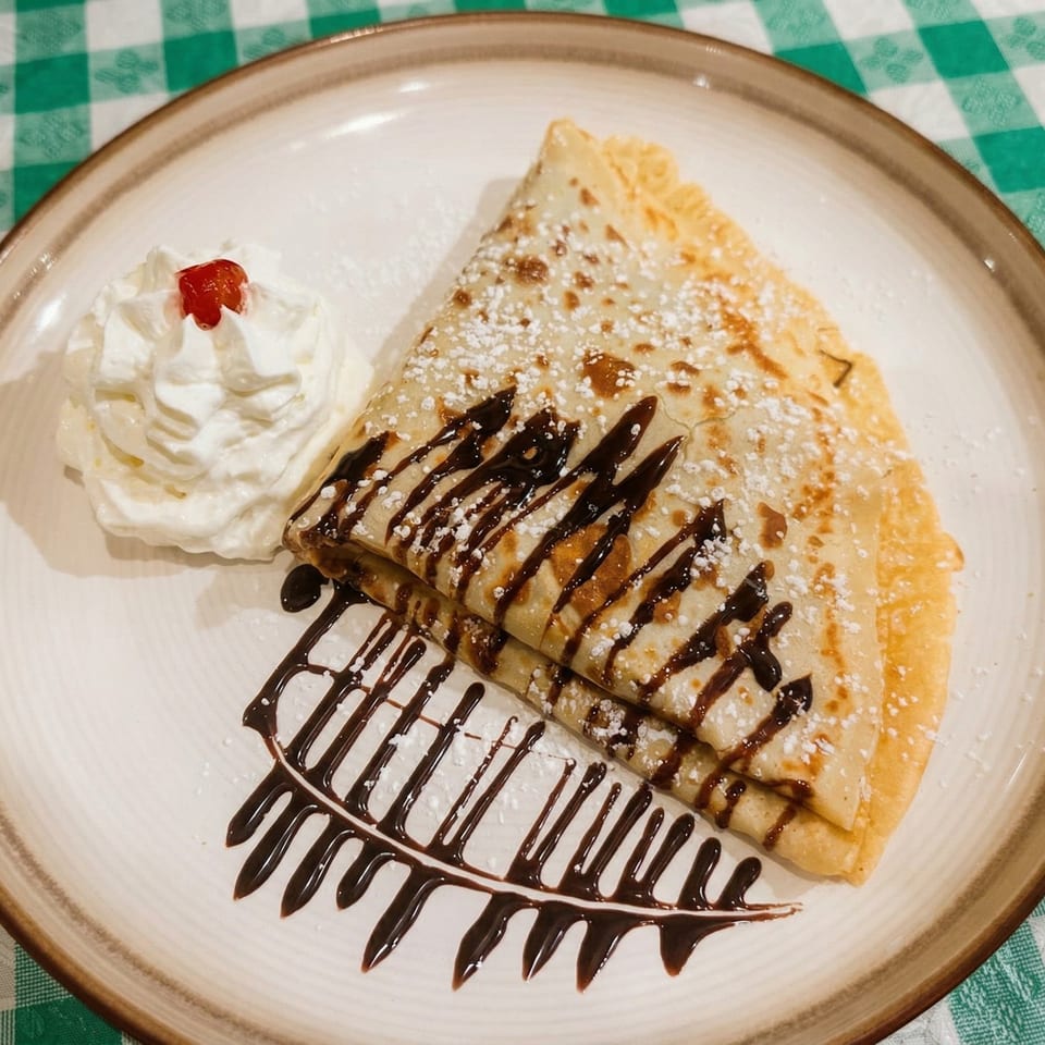 A folded crepe with chocolate drizzle, powdered sugar, whipped cream, and a cherry on a white plate.